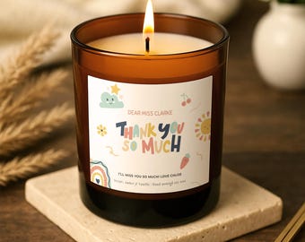 Puede incluir: Una vela de cera de soja encendida en un frasco de vidrio marrón con una etiqueta blanca que incluye el texto "Dear Miss Clarke", "Thank you so much" y "I'll miss you so much! Love Chloe". La etiqueta muestra ilustraciones coloridas.