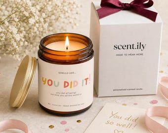 May include: A lit soy wax candle in an amber glass jar with a label reading "SMELLS LIKE... YOU DID IT! you did amazing! we love you all the world xx". It includes a white gift box with a dark red ribbon and a card reading "You did so well!"