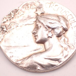 K&ouml;nnte beinhalten: Silberfarbene runde Medaille mit dem Profil einer Frau mit flie&szlig;endem Haar und einem sternf&ouml;rmigen Ohrring. Der Rand der Medaille tr&auml;gt die Inschrift "CREPUSCUL E Aurora". Der Hintergrund zeigt eine Landschaft mit Flederm&auml;usen.