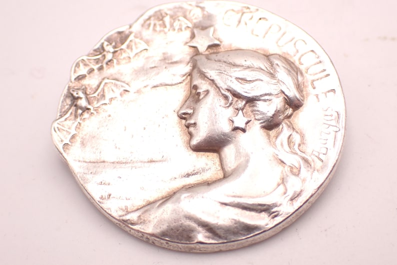 K&ouml;nnte beinhalten: Silberfarbene runde Medaille mit dem Profil einer Frau mit flie&szlig;endem Haar und einem sternf&ouml;rmigen Ohrring. Der Rand der Medaille tr&auml;gt die Inschrift "CREPUSCUL E Aurora". Der Hintergrund zeigt eine Landschaft mit Flederm&auml;usen.