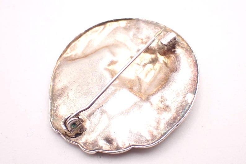 K&ouml;nnte beinhalten: Silberfarbene Brosche mit Nadelverschluss. Die Brosche hat eine ovale Form mit strukturierter Oberfl&auml;che und dekorativem Rand. Die Nadel ist auf der R&uuml;ckseite der Brosche befestigt.