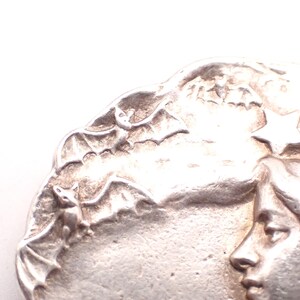 K&ouml;nnte beinhalten: Silberfarbenes Metallrelief mit Frauenprofil, Flederm&auml;usen und einem Stern. Das detaillierte Design zeigt ein Frauengesicht, Flederm&auml;use im Flug und einen Stern vor einem strukturierten Hintergrund. Nahaufnahme eines dekorativen Elements.