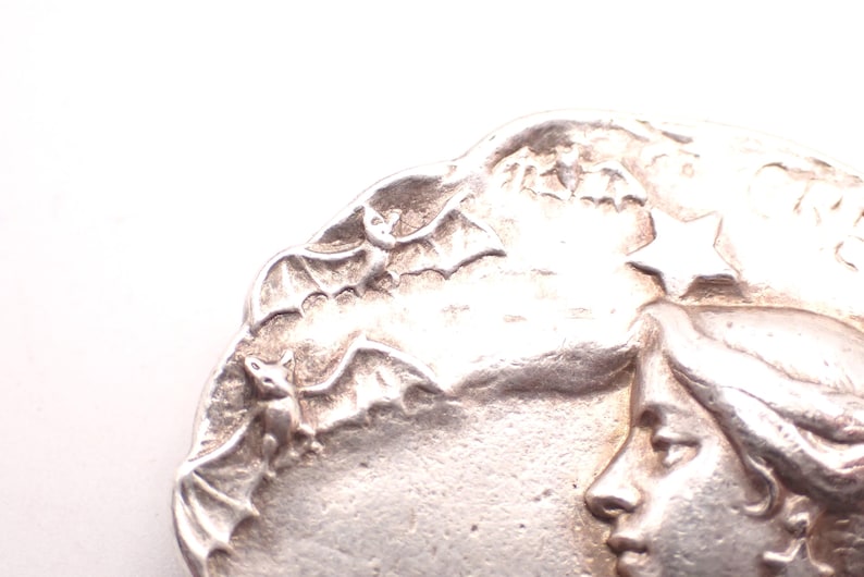 K&ouml;nnte beinhalten: Silberfarbenes Metallrelief mit Frauenprofil, Flederm&auml;usen und einem Stern. Das detaillierte Design zeigt ein Frauengesicht, Flederm&auml;use im Flug und einen Stern vor einem strukturierten Hintergrund. Nahaufnahme eines dekorativen Elements.