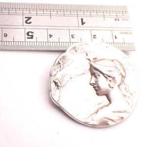 K&ouml;nnte beinhalten: Ein silberfarbener, runder Anh&auml;nger mit dem Profil eines Frauengesichts. Der Anh&auml;nger hat einen Durchmesser von etwa 3,8 cm. Ein Metalllineal liegt zur Gr&ouml;&szlig;enbestimmung dar&uuml;ber.
