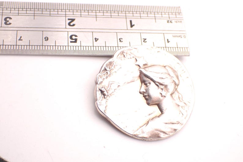 K&ouml;nnte beinhalten: Ein silberfarbener, runder Anh&auml;nger mit dem Profil eines Frauengesichts. Der Anh&auml;nger hat einen Durchmesser von etwa 3,8 cm. Ein Metalllineal liegt zur Gr&ouml;&szlig;enbestimmung dar&uuml;ber.