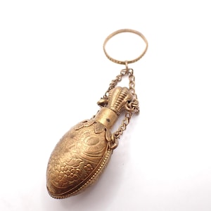 May include: A vintage perfume bottle in a gold tone, featuring detailed floral and geometric engravings. The oval-shaped bottle hangs from a chain and a ring. The design suggests a historical or antique style.