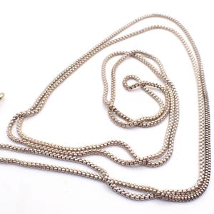 Puede incluir: Un collar plateado con un diseño de cadena delicado y tejido. El collar se muestra sobre un fondo blanco, resaltando su intrincada textura y el cierre al final. La cadena mide aproximadamente 76 cm de largo.