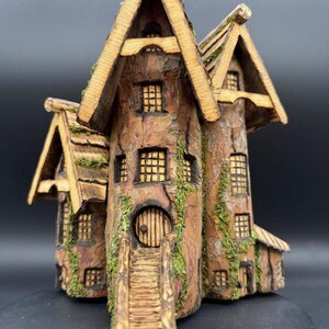 May include: A detailed, miniature wooden house with a rustic, bark-like exterior. The house features multiple windows, a round door, and a small staircase leading to the entrance. The roof is covered with a textured, moss-like material, and the overall design evokes a fairytale aesthetic.