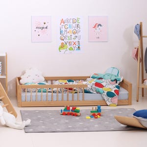 May include: A child's bedroom scene featuring a wooden bed with a blue mattress and colorful bedding. Decorative posters with the words "magical", "ABC", and "cute" hang on the wall. A gray rug with white polka dots and wooden toy train set are on the floor.