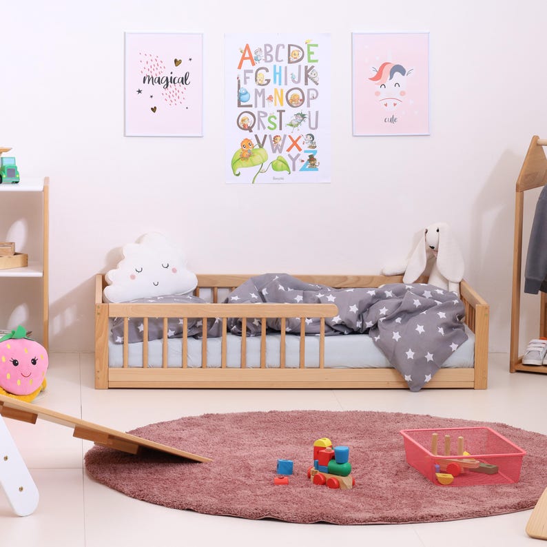 May include: A child's bedroom scene featuring a wooden bed with a gray star-patterned comforter and a cloud-shaped pillow. The room includes a pink rug, colorful toys, and decorative posters with the words "magical" and "cute".