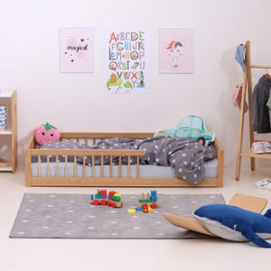 May include: A light wood toddler bed with a grey and white star patterned bedding. The bed has a safety rail on one side. There is a grey rug with white polka dots on the floor. The rug has a wooden toy whale and a toy train on it. There are three posters on the wall: one with the alphabet, one with a unicorn, and one with the word "magical".