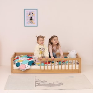 May include: Two young girls are playing on a light wood toddler bed with a colorful balloon-themed blanket. A framed Minnie Mouse print hangs on the wall. A plush white seal toy sits on the bed.