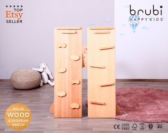 May include: Two wooden climbing structures for children, one with a ladder and the other with climbing holds. The structures are made of solid European beech wood and are freestanding. The structures are on a pink rug in front of a light gray wall. The text "brubi HAPPY KIDS" is on the wall.