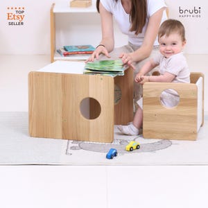Pode incluir: Conjunto de mesa e cadeira infantil em madeira com acabamento natural. A mesa tem um tampo branco e um orifício circular na lateral. Uma criança está sentada numa cadeira a condizer. Dois carros de brinquedo estão no tapete.