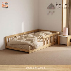 Op de afbeelding: Een peuterbed van massief eikenhout met veiligheidsrails. Het bedframe is van natuurlijk hout, gecombineerd met een beige dekbed en kussen. De kamer heeft een neutrale kleurstelling. Het bed is ongeveer 140 cm lang.