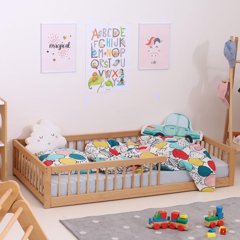 May include: A wooden toddler bed with a light blue mattress, colorful bedding, and decorative pillows shaped like a cloud and a car. The room features framed artwork and a wooden toy train on a gray rug.