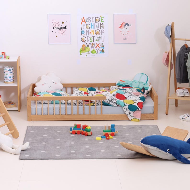May include: A child's bedroom with a wooden bed, a gray rug with white polka dots, and colorful bedding. The room is decorated with framed artwork and a wooden clothing rack. Toys, including a train and a whale, are scattered on the rug.