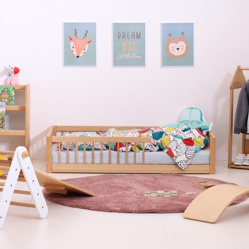 May include: A light wood toddler bed with a colorful patterned blanket and pillow. Three framed art prints hang above the bed. A pink rug and wooden toys are on the floor. The text "Dream Big Little One" is on one of the prints.