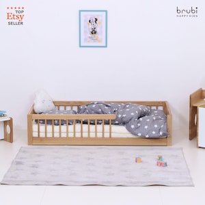 Könnte beinhalten: Ein Holzkinderbett mit einer grauen Bettdecke und einem Kissen mit Sternenmuster. Auf dem Boden liegt ein grauer Teppich mit Sternenmuster. Ein gerahmter Minnie-Maus-Druck hängt an der Wand. Das Bett hat ein Sicherheitsgeländer.