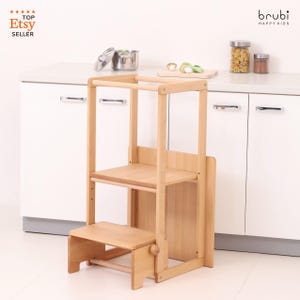 May include: A wooden step stool with a safety rail and a lower step. The stool is light brown and has a smooth finish. It is designed to help children reach higher surfaces in the kitchen.