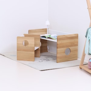 Pode incluir: Uma secretária infantil de madeira clara com duas cadeiras anexas. A secretária tem um tampo branco e uma pequena lâmpada. Um livro está aberto na secretária. As cadeiras têm recortes circulares. A secretária está sobre um tapete claro.