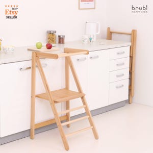 Può includere: Uno sgabello di aiuto per la cucina in legno chiaro con una piccola piattaforma e gradini a forma di scala. Lo sgabello ha un piccolo tavolo in cima. È visibile anche un piano di lavoro bianco con un tagliere, mele e barattoli. Il logo del venditore Etsy è visibile.