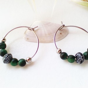 Peut inclure: Une paire de boucles d'oreilles créoles argentées avec des perles vertes et noires. Les perles ont un motif en spirale blanc.