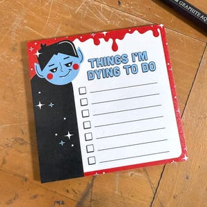 May include: A notepad with a red border and the text "THINGS I'M DYING TO DO" in white. The notepad features a cartoon vampire character and a checklist. A black graphite pencil is in the background.