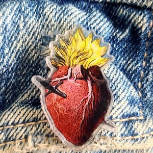 Könnte beinhalten: Eine durchsichtige Plastiknadel mit einem detaillierten Bild eines roten anatomischen Herzens mit einer gelben Flamme oben. Das Herz hat eine sichtbare Venenstruktur und einen einzigen schwarzen Pfeil, der es durchbohrt.