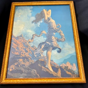 May include: Framed print of a woman in a flowing dress with her arms raised, set against a blue sky with clouds and mountains. The artwork is in a gold-colored frame.