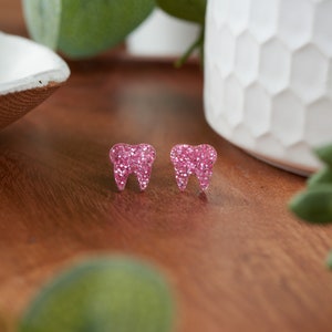 May include: A pair of pink glitter tooth-shaped earrings. The earrings are small and delicate, and they would make a great gift for a dentist or someone who loves teeth.