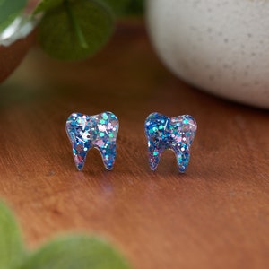May include: A pair of tooth-shaped earrings with blue, pink, and iridescent glitter embedded in the clear resin.
