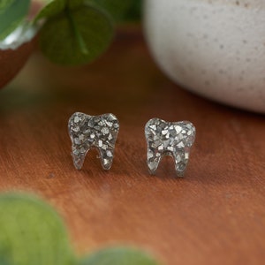 May include: A pair of silver glitter tooth stud earrings. The earrings are made of clear resin with silver glitter embedded inside.