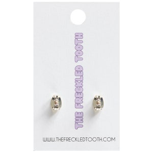 Puede incluir: Un par de pendientes de plata con un peque&ntilde;o cristal transparente en el centro. Los pendientes est&aacute;n en una tarjeta blanca con el texto "THE FRECKLED TOOTH" y "WWW.THEFRECKLEDTOOTH.COM".