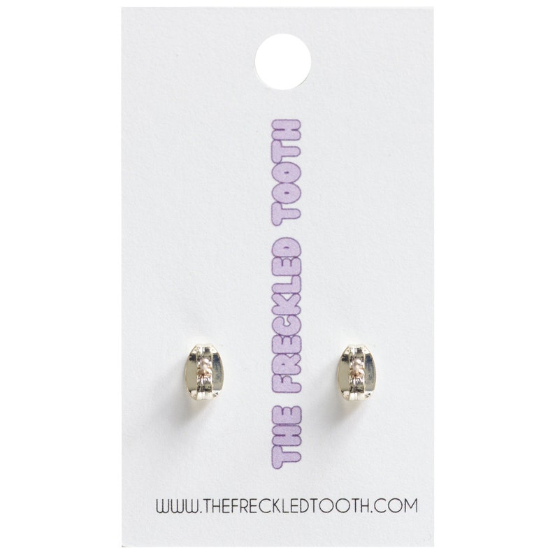 Puede incluir: Un par de pendientes de plata con un peque&ntilde;o cristal transparente en el centro. Los pendientes est&aacute;n en una tarjeta blanca con el texto "THE FRECKLED TOOTH" y "WWW.THEFRECKLEDTOOTH.COM".