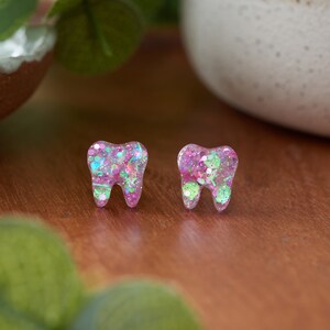 May include: A pair of pink and green glitter tooth earrings. The earrings are shaped like teeth and are covered in iridescent glitter.