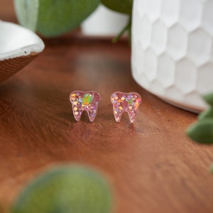 May include: A pair of pink tooth-shaped earrings with iridescent glitter embedded inside.