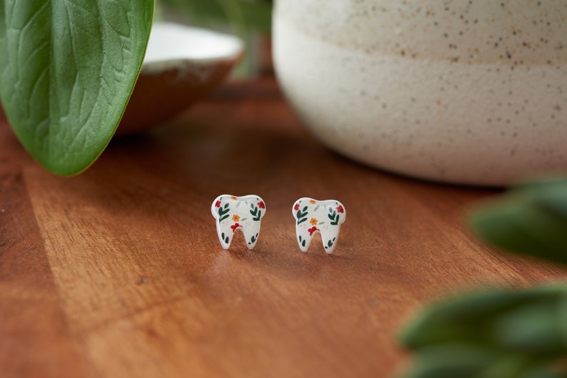 Puede incluir: Un par de pendientes blancos en forma de diente con un patr&oacute;n floral en tonos rojos, naranjas, verdes y blancos.