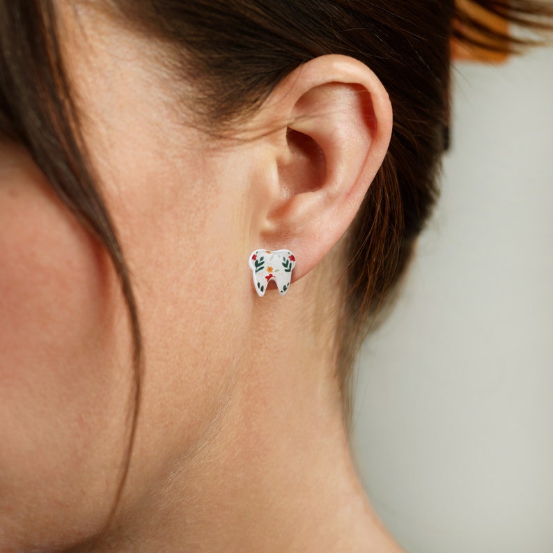 Puede incluir: Un par de pendientes blancos en forma de diente con un dise&ntilde;o floral en tonos verdes, rojos y amarillos.