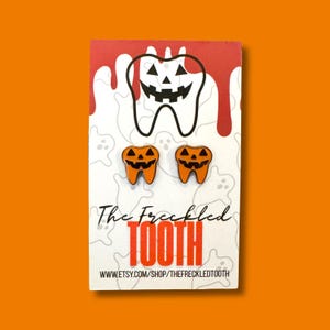May include: A pair of orange pumpkin-shaped earrings with black carved faces, displayed on a card with a tooth graphic and the text "The Freckled TOOTH". The background is orange.