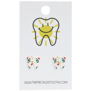 Puede incluir: Un par de pendientes blancos en forma de diente con un patr&oacute;n floral. Los pendientes est&aacute;n en una tarjeta blanca con un gr&aacute;fico de diente sonriente y el texto "www.thefreckledtooth.com".