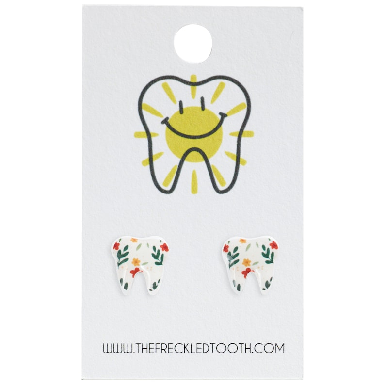 Puede incluir: Un par de pendientes blancos en forma de diente con un patr&oacute;n floral. Los pendientes est&aacute;n en una tarjeta blanca con un gr&aacute;fico de diente sonriente y el texto "www.thefreckledtooth.com".