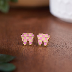 May include: A pair of gold-tone earrings shaped like teeth with a pink and yellow checkered pattern.