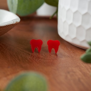 May include: A pair of red tooth-shaped earrings. The earrings are made of a smooth, glossy material and are a fun and quirky accessory.