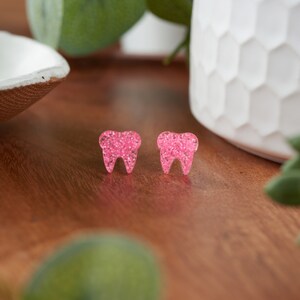 May include: A pair of pink glitter tooth earrings. The earrings are small and have a sparkly finish.