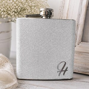 May include: A silver glitter-finished metal flask with a rectangular shape and rounded corners. The flask features a polished silver screw-top cap and a dark engraved letter H on the lower right side of the front surface of the container.