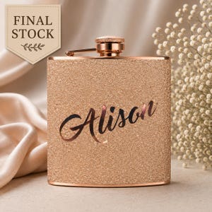 Puede incluir: Una petaca de purpurina en oro rosa con tapa de rosca metálica. La superficie frontal presenta el nombre Alison en una caligrafía oscura. Una etiqueta beige en la esquina superior izquierda muestra el texto FINAL STOCK con una hoja.