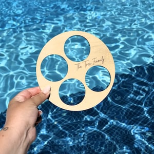 May include: A circular wooden drinks holder with five holes, held against a blue water background. The text "The Tree Family" is engraved on the wood. The holder is made of light-colored wood.