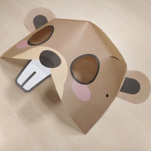 May include: A brown paper mask with black ears and eyes, a white nose and teeth, and pink cheeks. The mask is designed to look like a beaver.