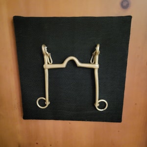 May include: A gold-colored metal horse bit with a curved mouthpiece and two rings on either side.